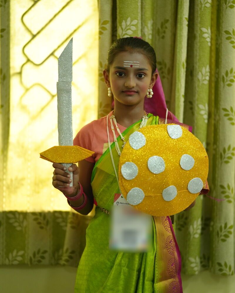 A girl in a sari holding a paper sword and a spotted shield